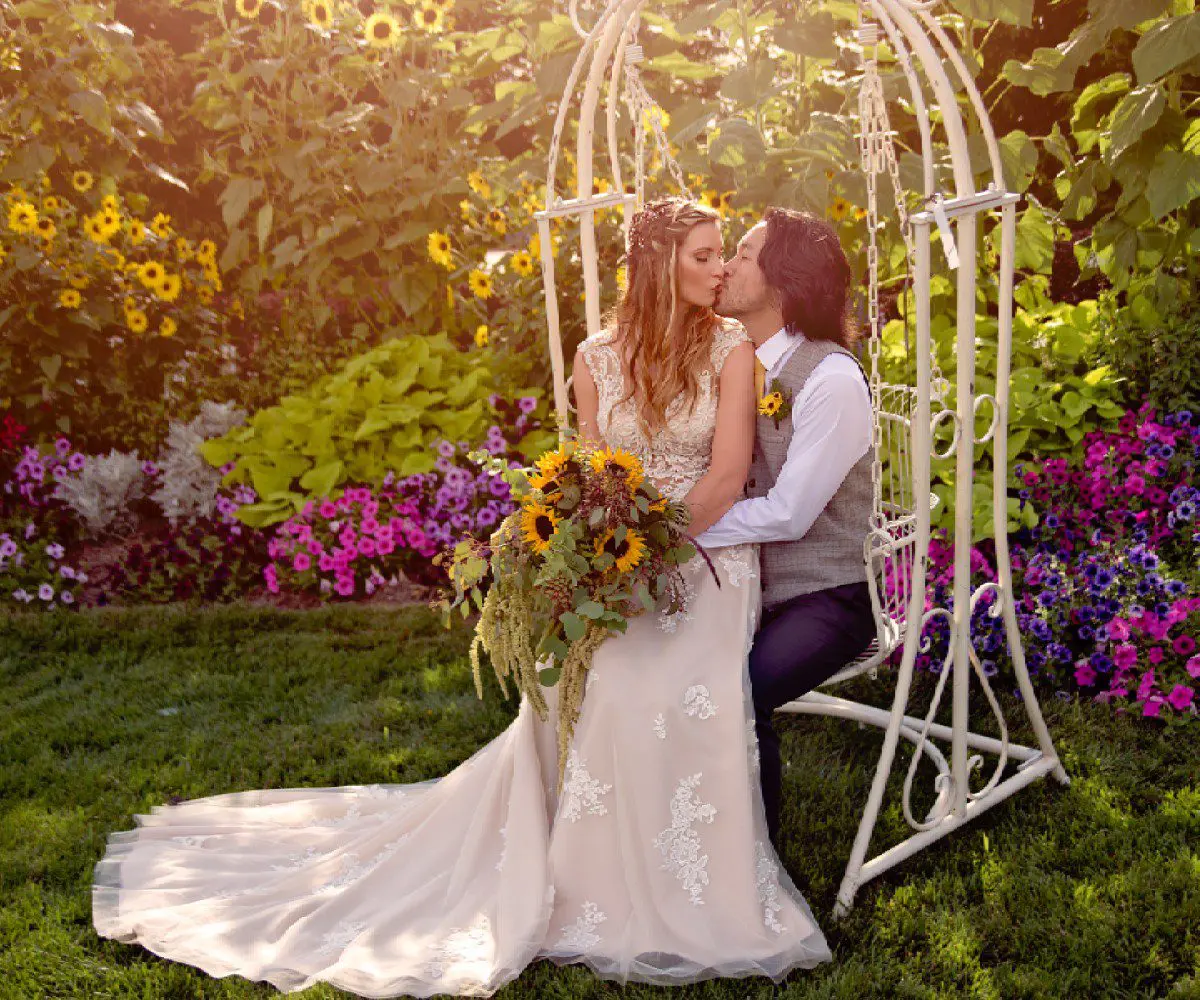 A couple kisses on a swing adorned with flowers, surrounded by a vibrant garden at sunset.