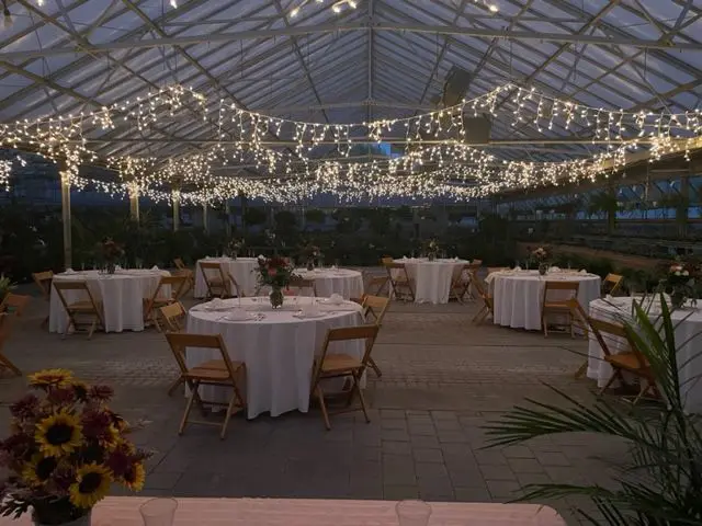 Evening view of an outdoor event space under a tent, decorated with string lights and set with round tables, each covered with a white cloth and floral centerpieces featuring an assortment of perennial flowers.