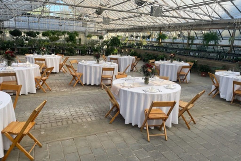An elegant event setup inside a greenhouse with round tables dressed in white, wooden chairs, and decorative centerpieces.