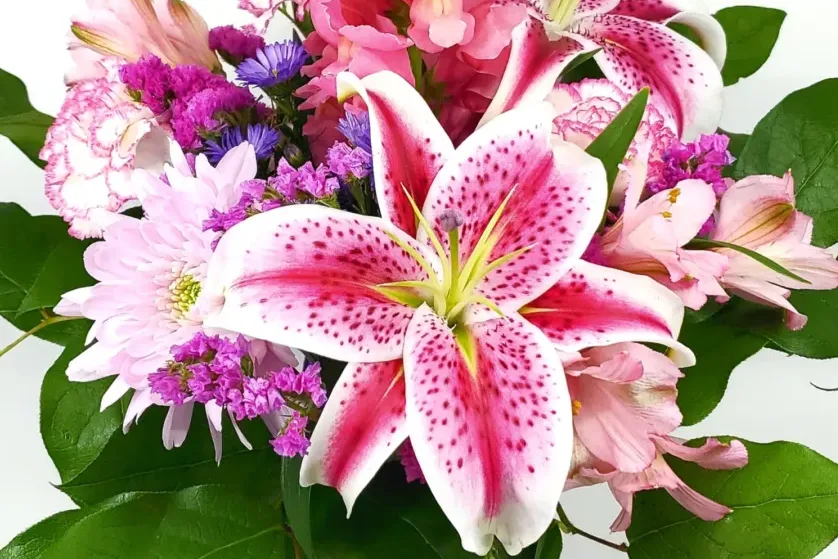 A vibrant bouquet featuring a prominent pink and white speckled lily, surrounded by pink and purple flowers and green leaves, crafted by a skilled florist.