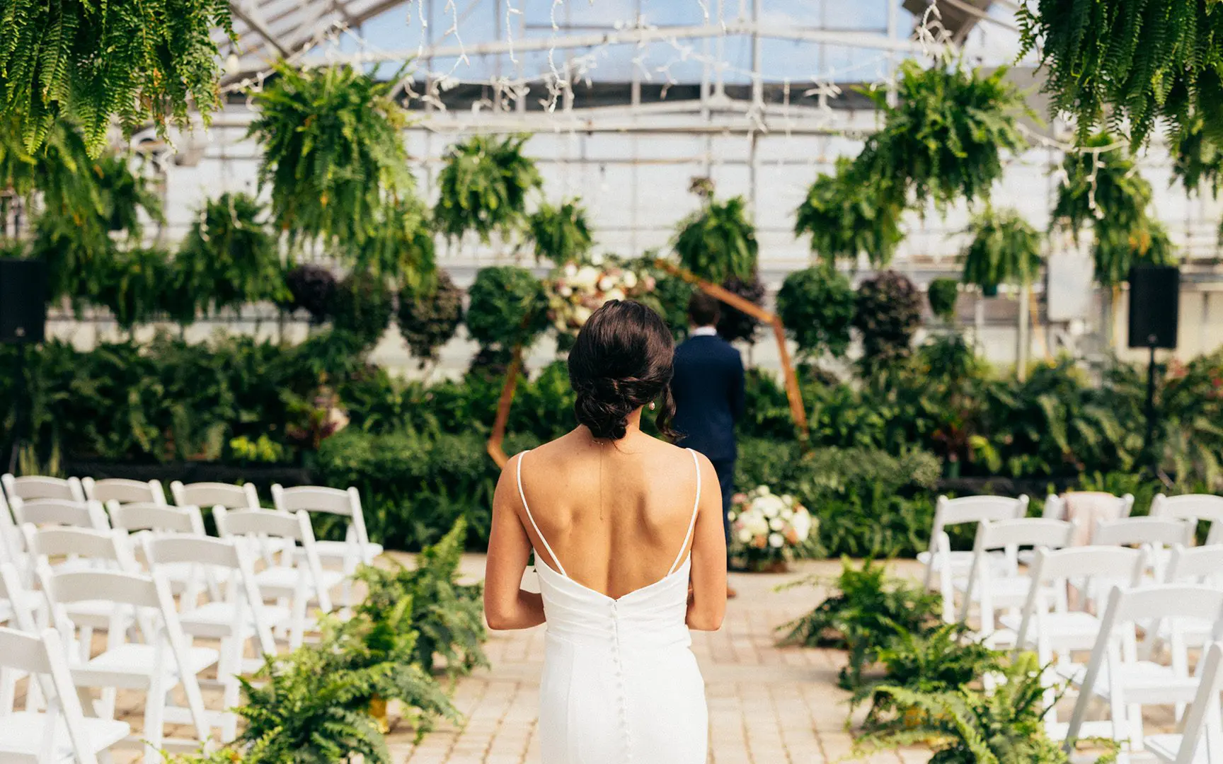 Bride in a white dress walking towards groom in a garden wedding ceremony setup with white chairs and hanging green plants.