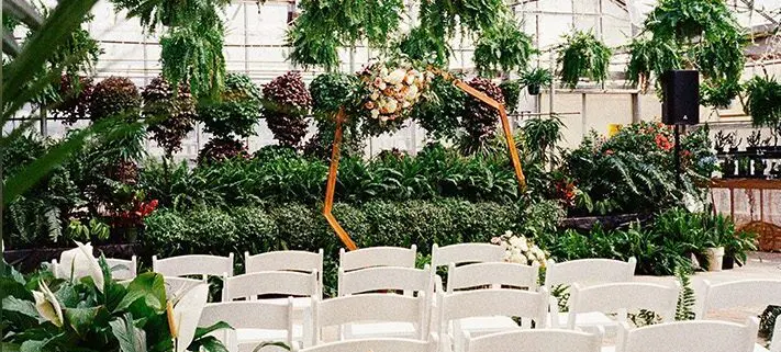 A wedding arch decorated with flowers, surrounded by rows of white chairs, set inside a lush greenhouse filled with various plants.
