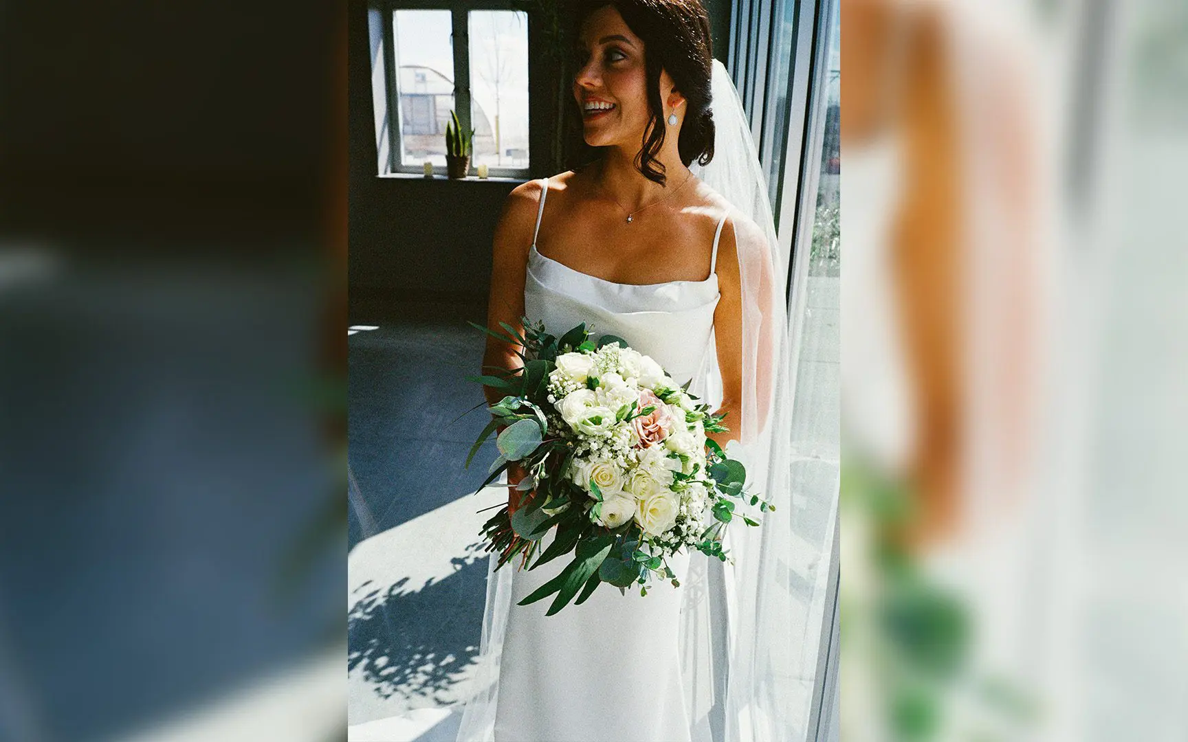 A bride in a white dress smiling and holding a bouquet of white flowers, standing indoors with natural light streaming in.