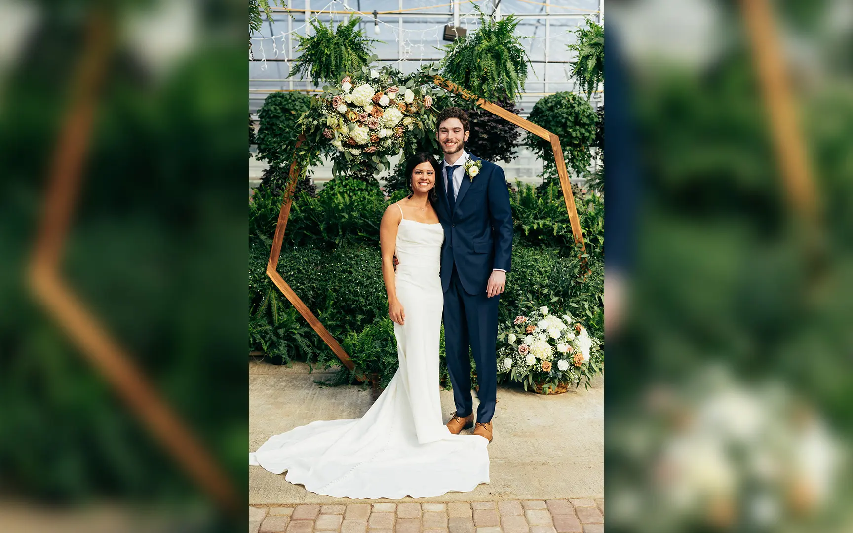 A newlywed couple posing under a floral arch in a greenhouse, the bride in a white dress and the groom in a blue suit.