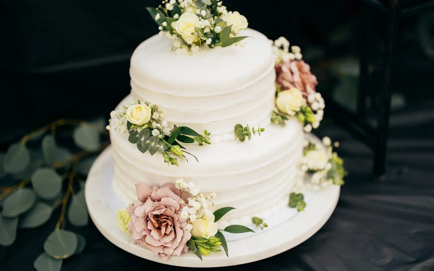 Three-tiered wedding cake adorned with creamy white icing and decorated with pink and yellow roses, displayed on a white stand.