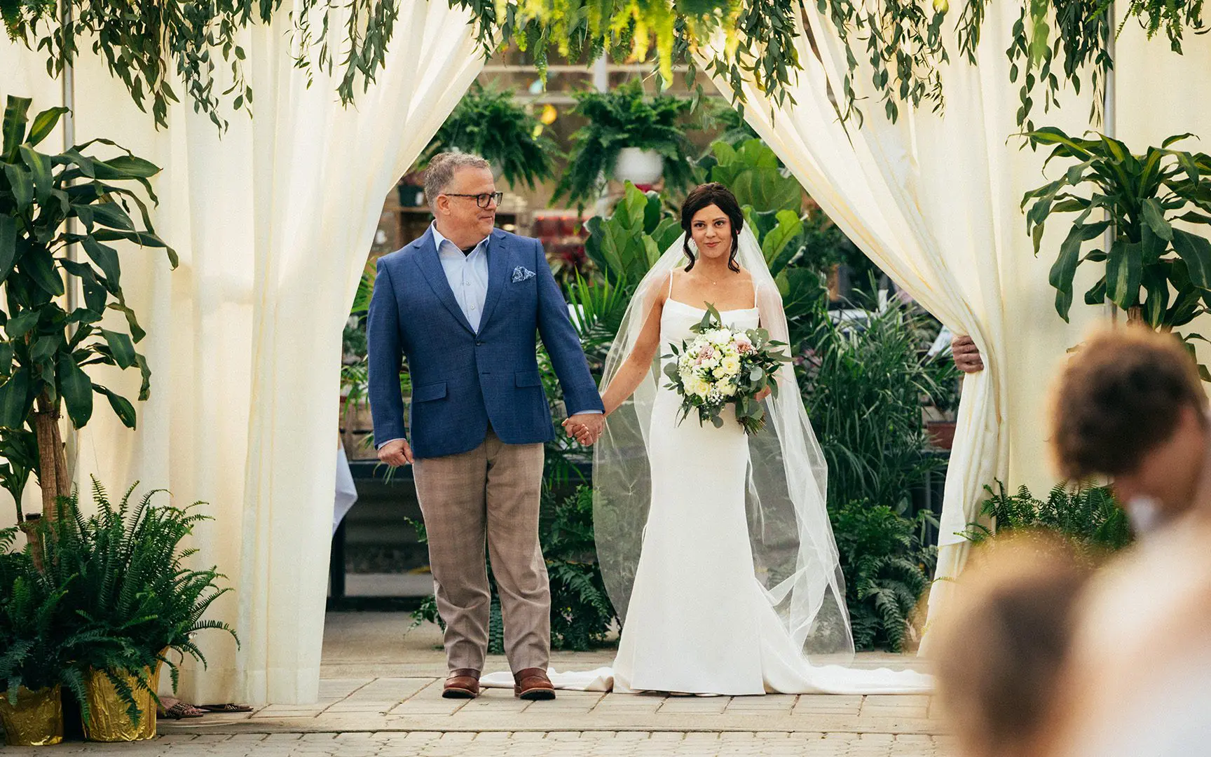 A bride in a white dress and her father walking down the aisle, surrounded by plants and draped curtains.