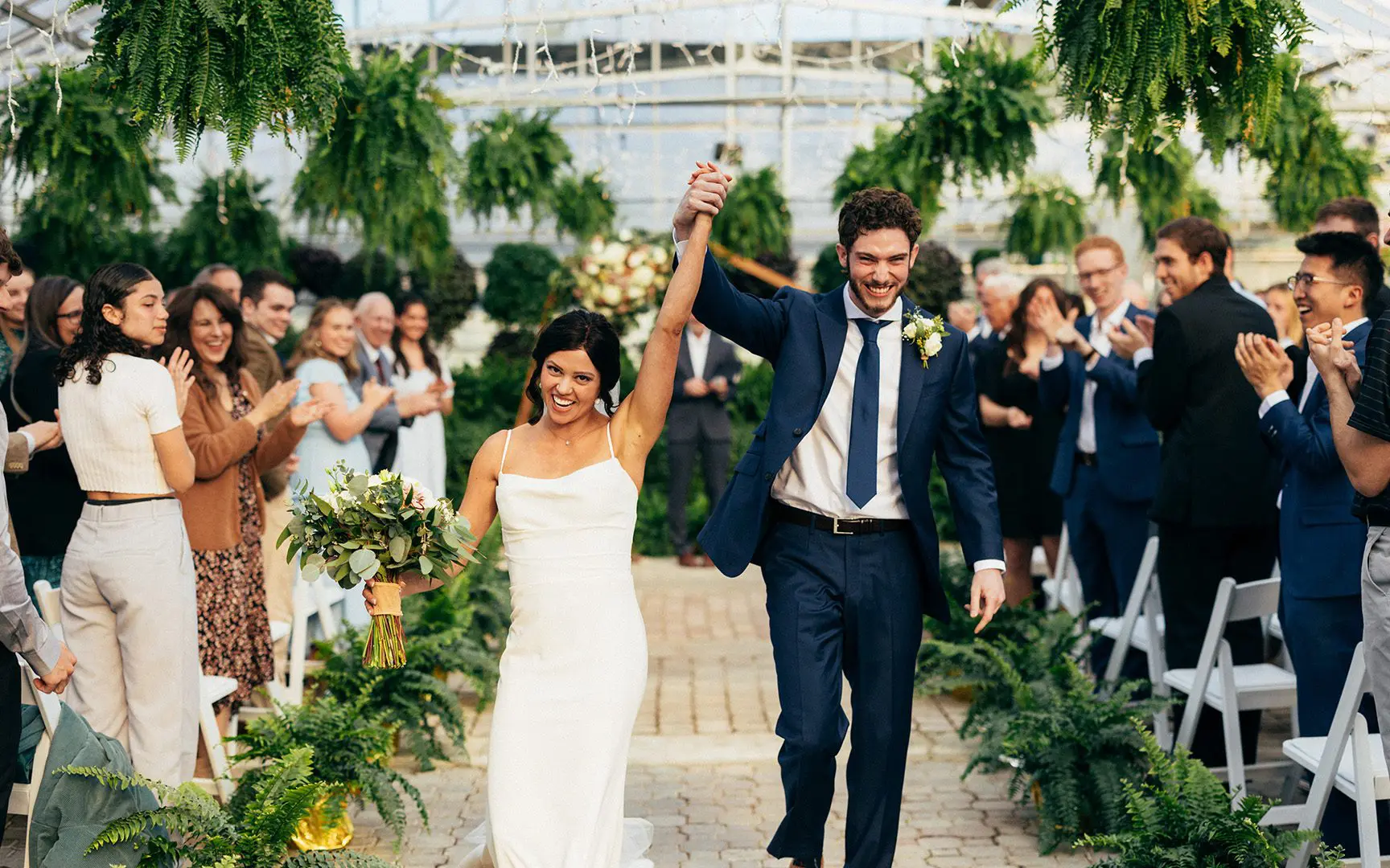 Newlywed couple joyfully walking down the aisle, holding hands and smiling, surrounded by applauding guests in a greenhouse setting.