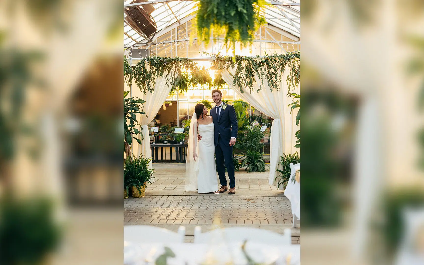A couple smiling together at a wedding venue decorated with hanging plants and white drapery, seen through a blurred floral archway.